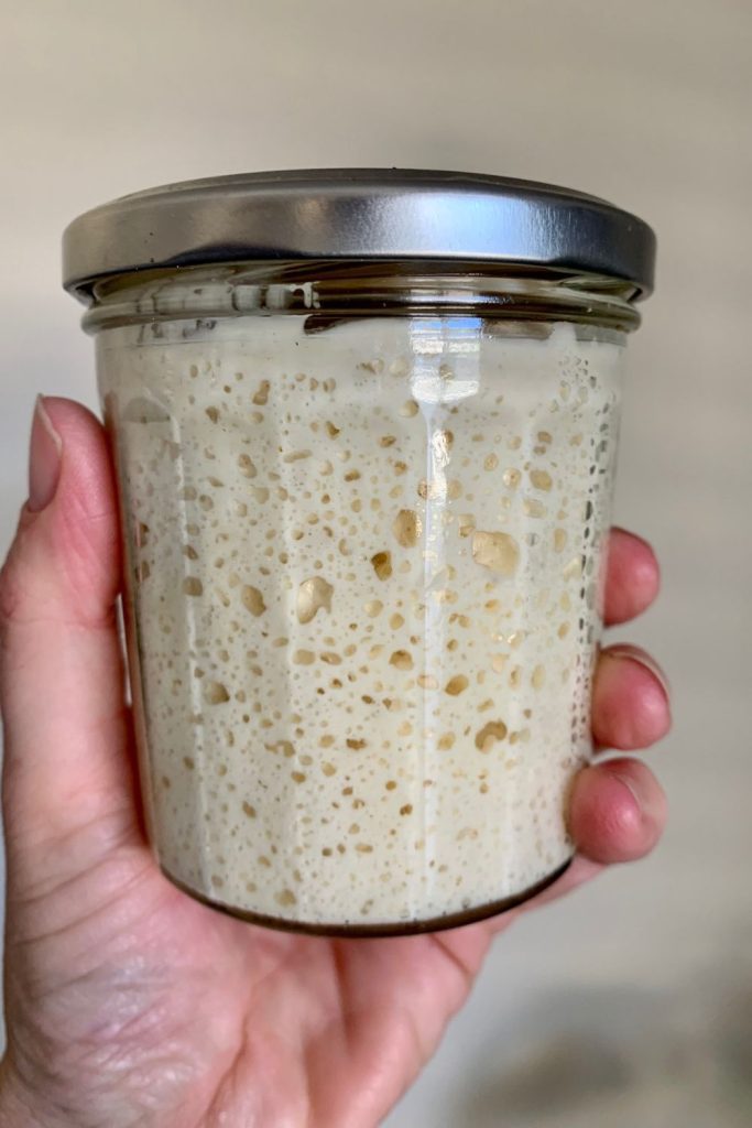 A small jar of sourdough starter with the lid on. The starter fills up most of the jar and is being held up for the photo.
