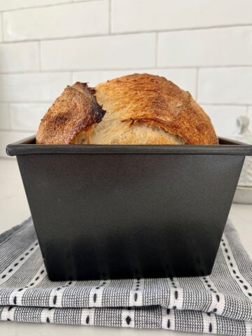 Loaf of sourdough bread that has been baked in a loaf pan sitting on a black and white dish towel in front of a white tiled wall.