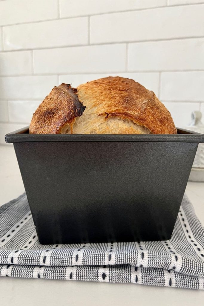 Loaf of sourdough bread that has been baked in a loaf pan sitting on a black and white dish towel in front of a white tiled wall.