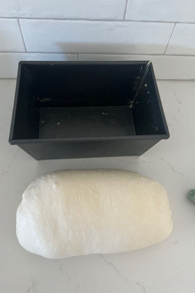 A loaf of sourdough that has been shaped into a loaf or batard shape sitting in front of a large loaf tin.