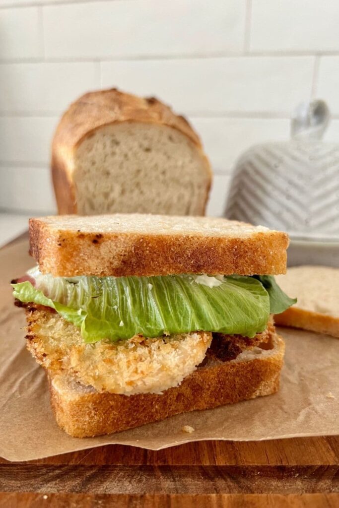 A loaf of sourdough bread baked in a loaf pan sitting on a wooden baord. There is a chicken and lettuce sandwich in the foreground and a butter dish in the background.