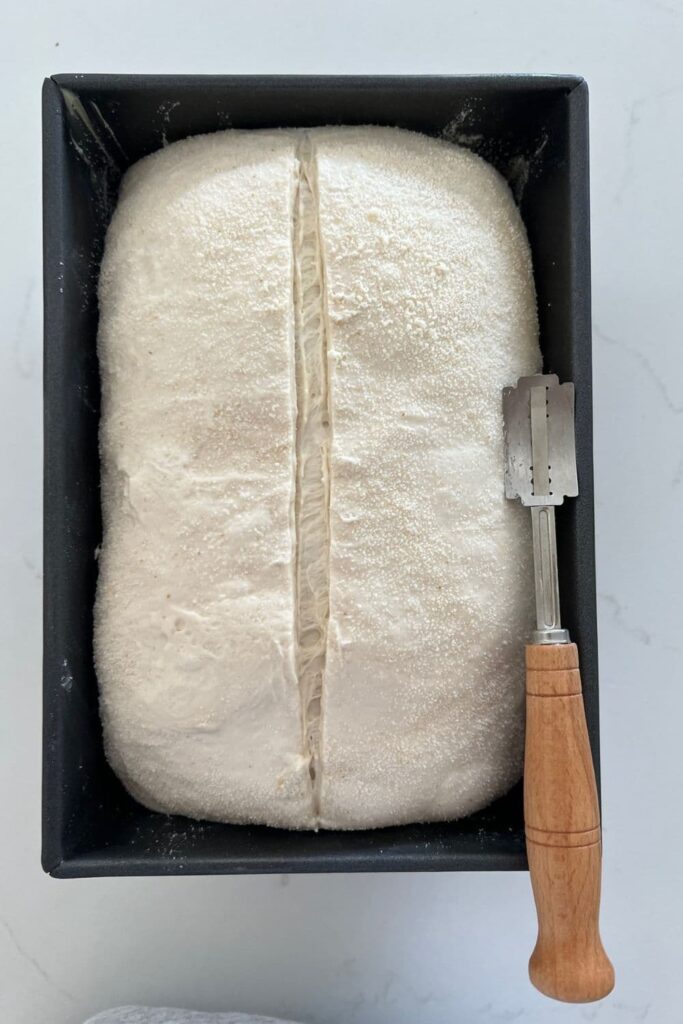 Loaf of sourdough bread in a loaf tin that has been scored down the centre. There is a wooden handled lame sitting on the edge of the bread tin.