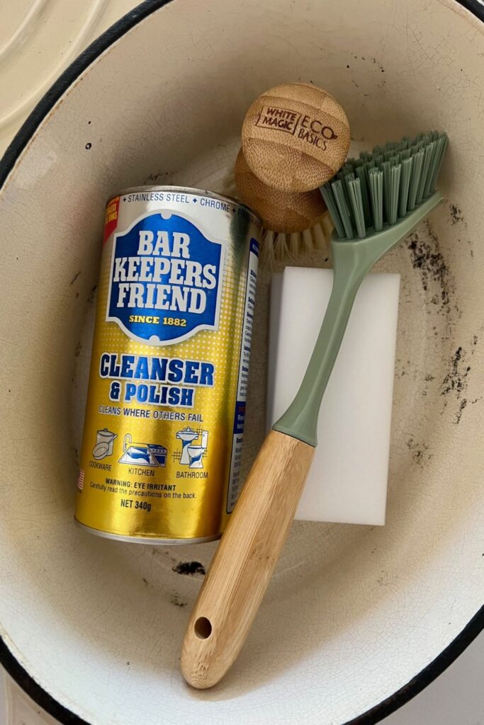 Cream enamel oval Dutch Oven with the lid off. There is a can of Bar Keeper's Friend sitting inside the Dutch Oven, along with a wooden pot brush, long handled plastic scrubbing brush and a magic eraser sponge.