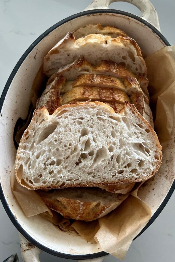 Loaf of long fermented sourdough bread sitting in a cream enamel Dutch Oven. There is a slice that has been placed on top of the loaf to show the crumb formation.