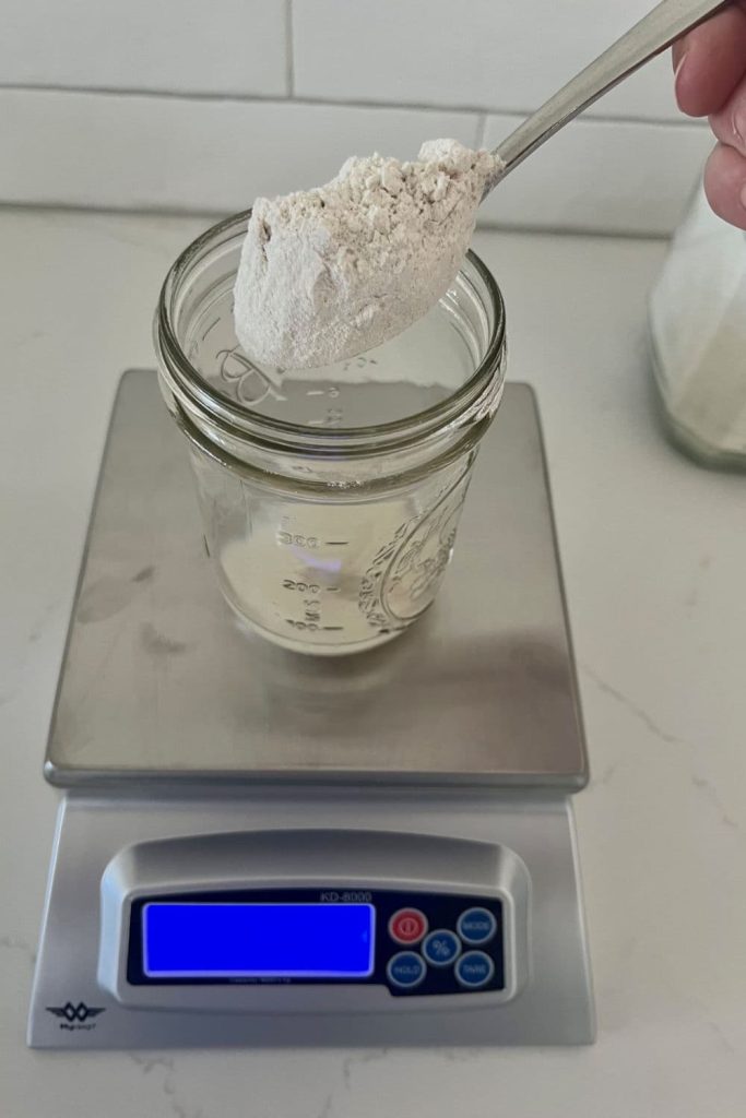 This photo shows a set of KD-8000 Baker's Scale with a small jar sitting on top. There is a spoon of rye flour being held over the opening of the jar.