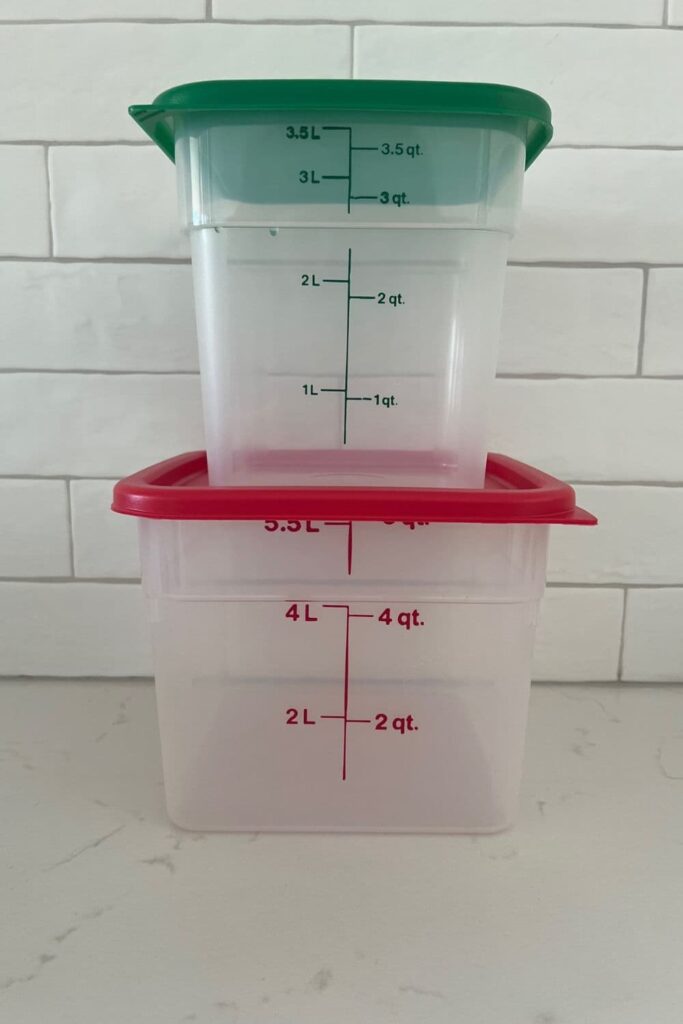 Two Cambro Containers stacked on top of each other. The one on the bottom has a red lid and is 6Q in size, the one on the top has. green lid and is 4Q in size. There is a white tiled wall in the background.