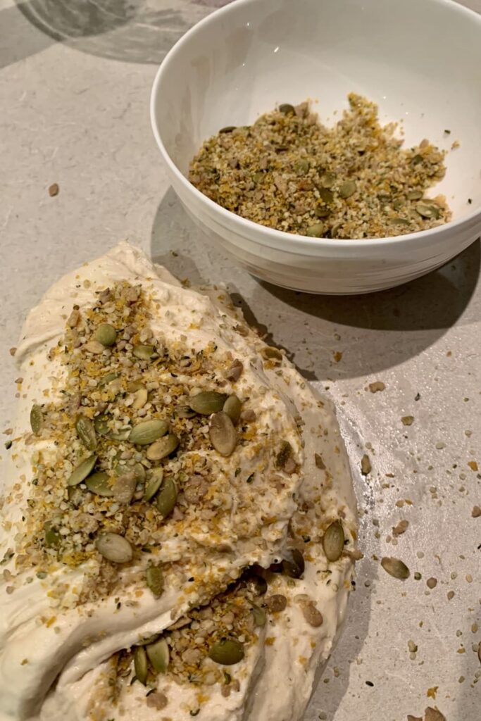 Multigrain sourdough bread dough sitting on the counter with soaked grains sprinkled through the dough. There is also a bowl of soaked grains sitting behind the dough.