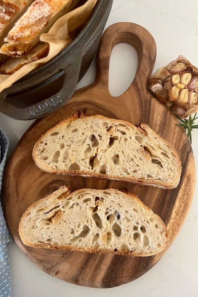 Roasted Garlic Sourdough Bread that has been sliced. There are 2 slices sitting on a round wooden board, as well as a head of roasted garlic in the background.