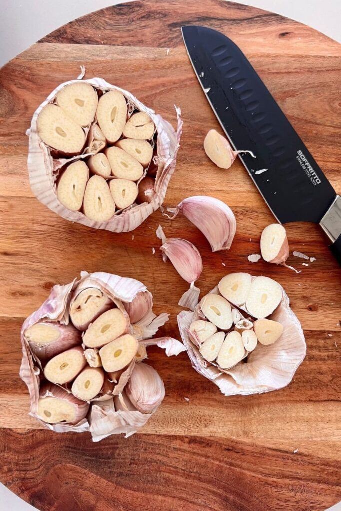 3 heads of garlic that have been sliced open to show the garlic cloves inside. They are sitting on a round wooden board and there is a black chef's knife to the right of the garlic.