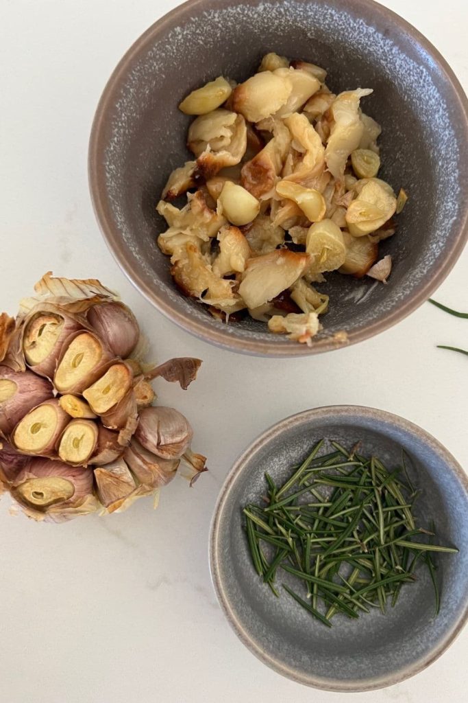 A head of roasted garlic sitting beside a stoneware bowl of fresh rosemary and another bowl of roasted garlic cloves that have been squeezed out of their skins.