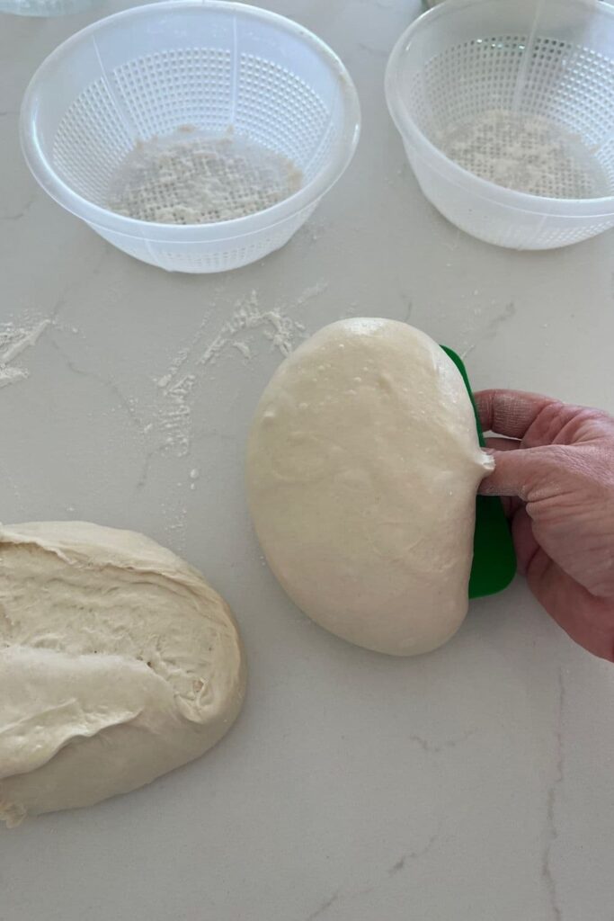 Two ricotta baskets sitting in the background with two dough balls being lifted by a green plastic dough scraper.