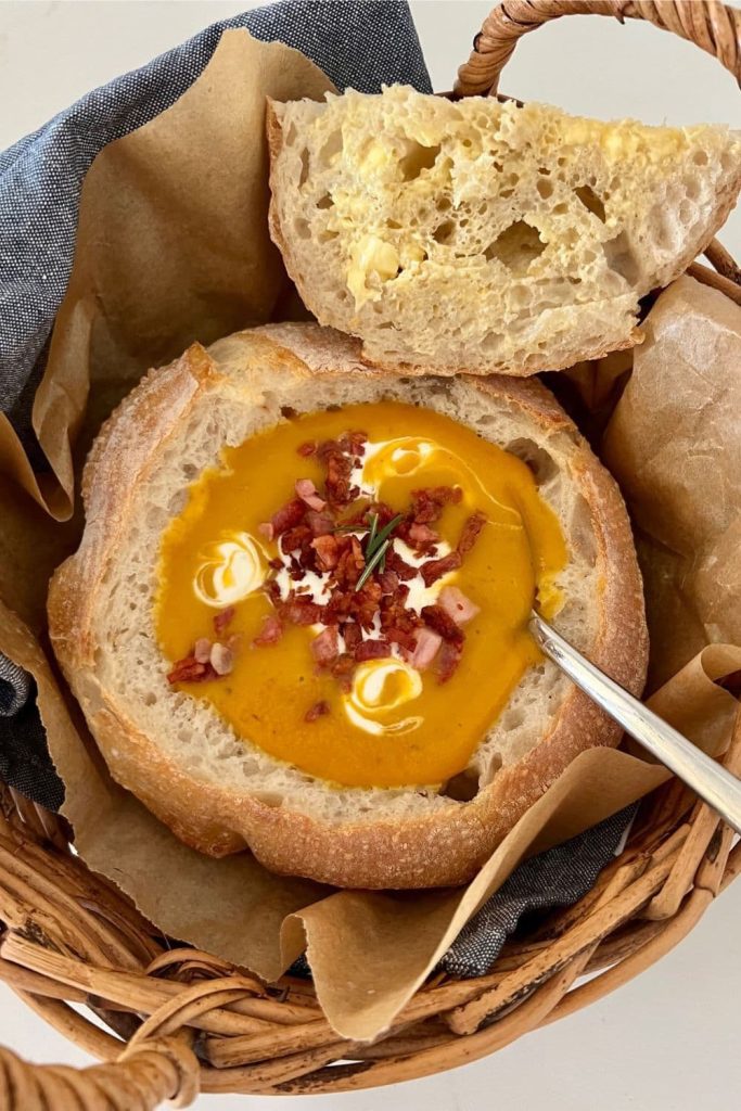 A sourdough bread bowl filled with orange pumpkin soup topped with sour cream and bacon. There is a piece of sourdough bread sitting alongside the bread bowl.