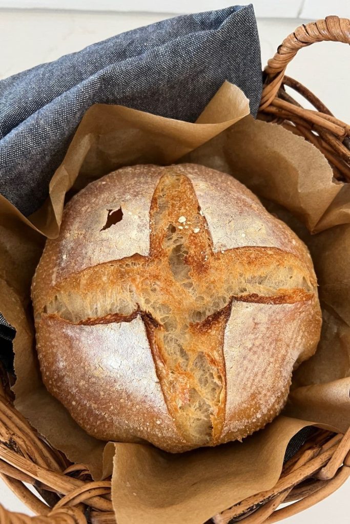 A sourdough bread bowl with a simple cross slashed into the top. It has been baked and is sitting inside a rattan basket lined with a denim blue dish towel.