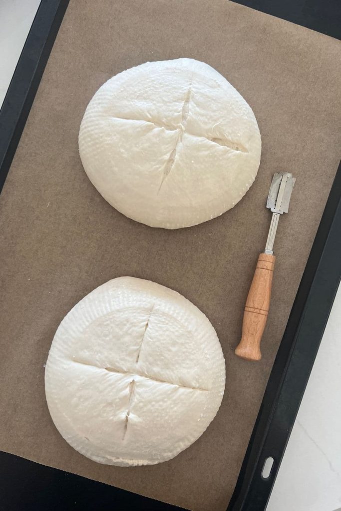 Two loaves of sourdough bread sitting on a piece of parchment paper. They have been scored with a cross on the top. There is a wooden handled lame sitting to the right of the bread bowls.