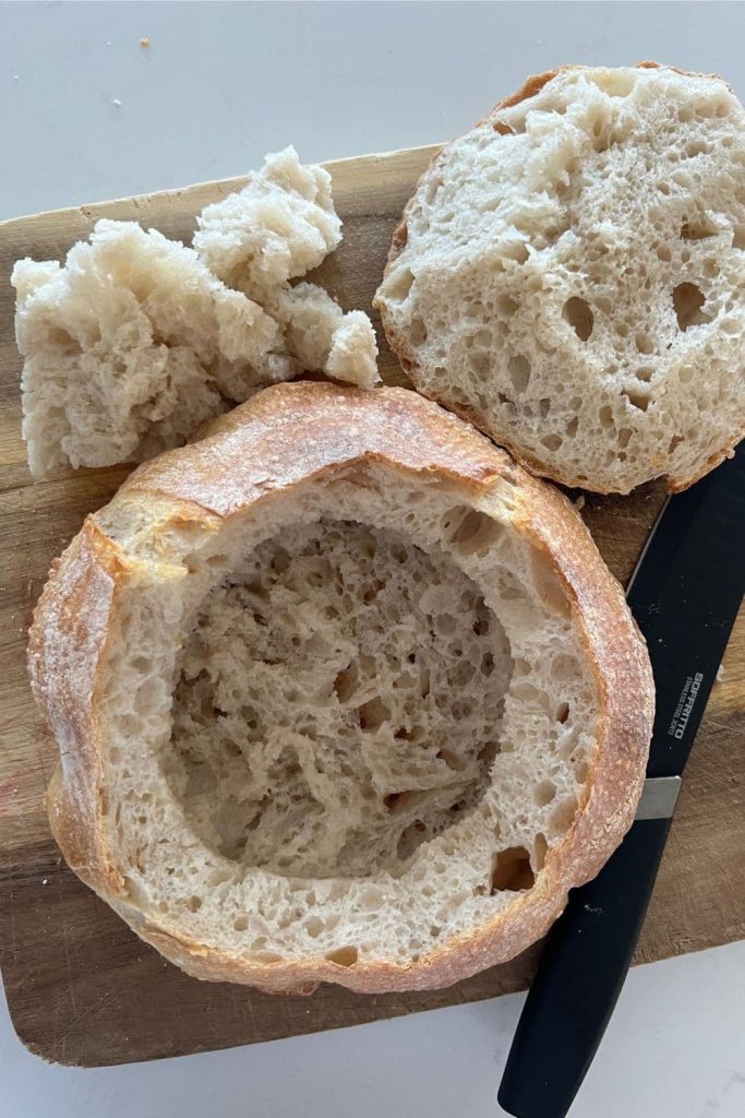 A sourdough bread bowl that has been hollowed out. There is a sharp, black knife sitting to the right of the bread bowl.