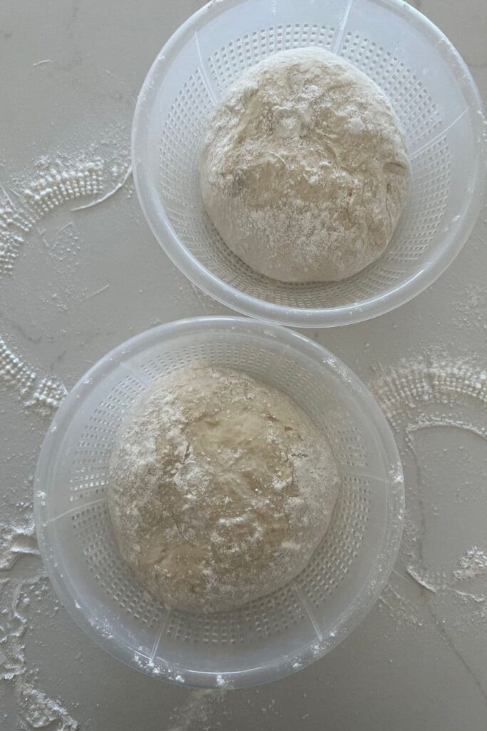 Two plastic ricotta baskets holding a loaf of sourdough each. There is a lot of rice flour sprinkled around them.