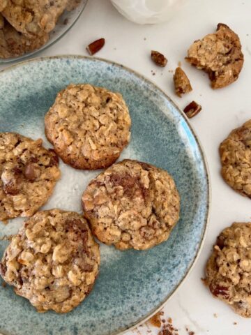 4 sourdough discard cowboy cookies on a blue stoneware plate. There is a glass of milk in the background and some cookie crumbs around the edges of the plate.