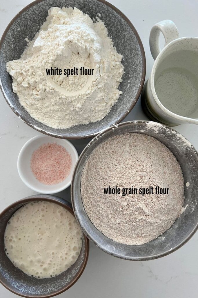 Ingredients for sourdough spelt bread. There is a bowl of white spelt flour, a bowl of whole grain spelt flour, bowl of salt, sourdough starter and jug of water.