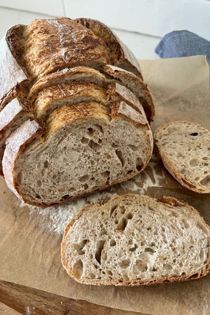 Loaf of sourdough spelt bread sitting on a wooden board. The loaf has been sliced into 5 slices. Two are laying on the board and 3 are sitting in front of the loaf of spelt sourdough. There is a blue dish towel in the backgroun.