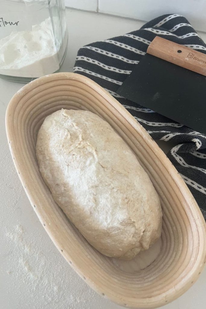 Loaf of sourdough that has been shaped into a batard and placed into an oval banneton. There is a jar of rice flour and a dough scraper in the photo as well.