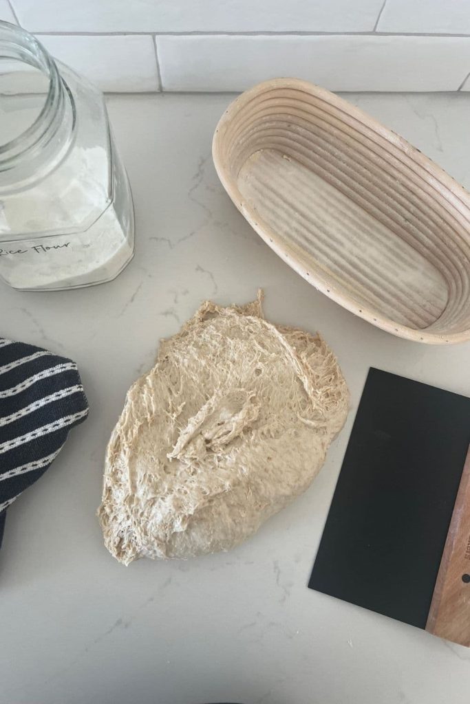 Spelt sourdough bread dough sitting on the kitchen counter. There is a glass jar of rice flour, black dough scraper, banneton and black and white dish towel.
