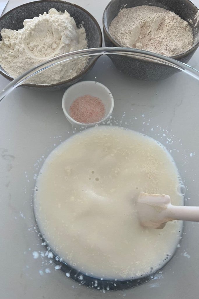 Glass bowl with water and sourdough starter being mixed together with a white jar spatula. There is a bowl of white and whole grain spelt flour and salt in the background.