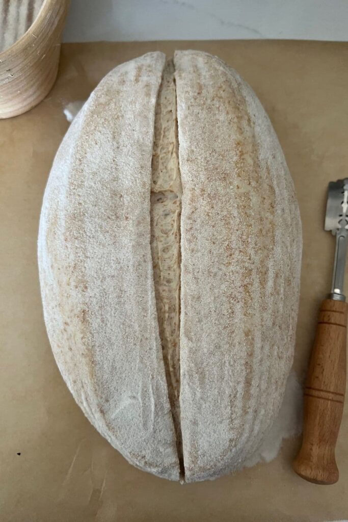 Sourdough batard that has been turned out of the banneton and placed on a piece of parchment paper. The dough has been scored in the centre with a lame which is lying next to the dough.