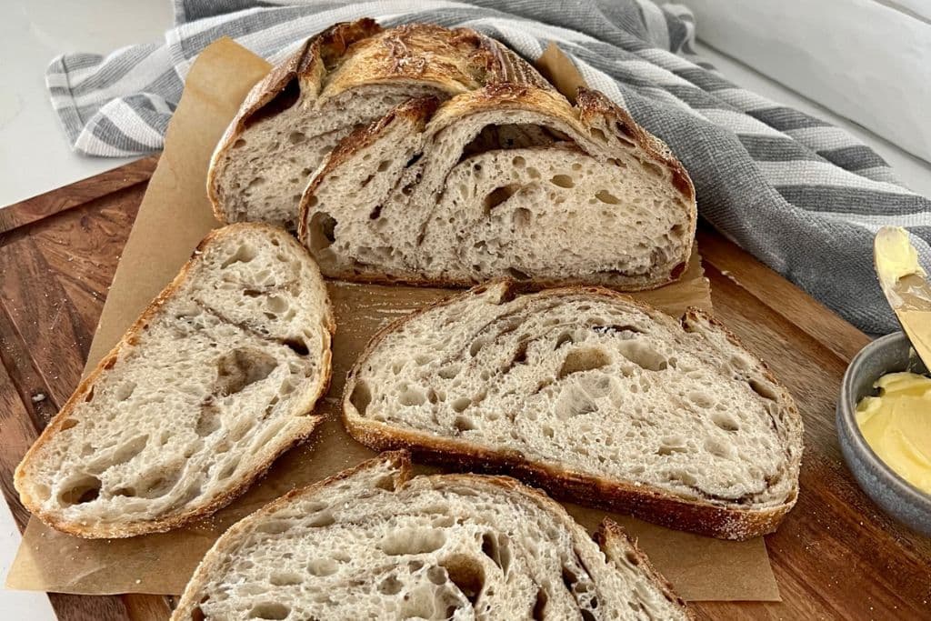 Cinnamon Swirl Sourdough Bread that has been sliced and is sitting on a bread board next to a small dish of butter. There is a grey and white striped dish towel in the background.