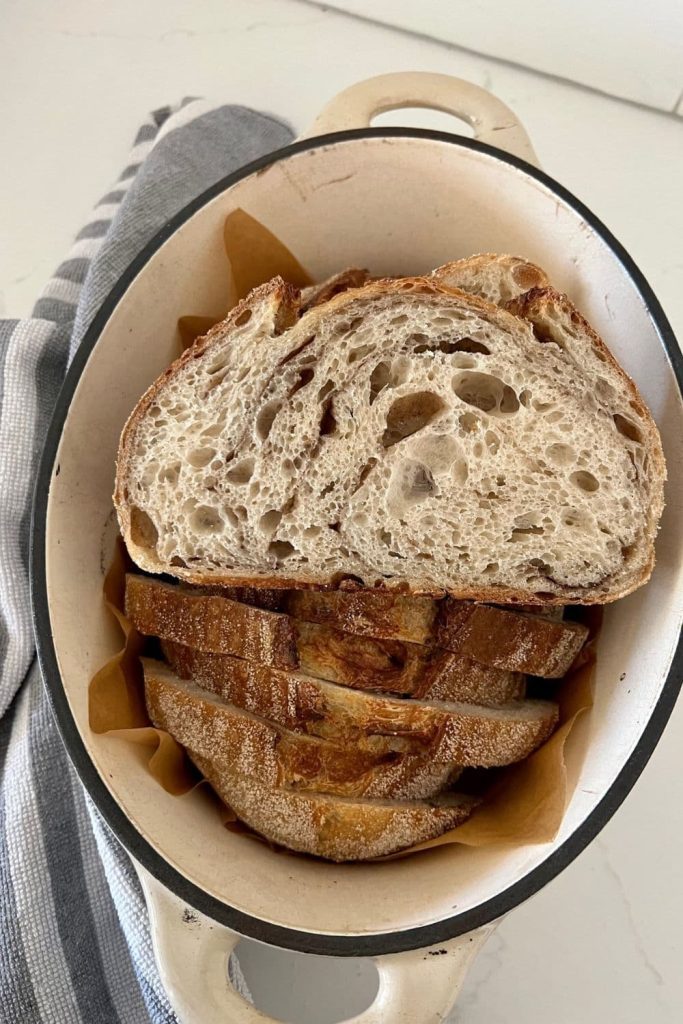 Loaf of sourdough cinnamon bread sitting in a cream colored enamel Dutch Oven. There is a slice of bread sitting on top of the loaf to show the cinnamon swirl and open crumb.