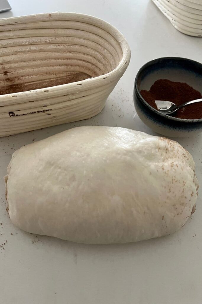 Batard shaped cinnamon swirl sourdough sitting in front of an oval shaped banneton that has been dusted with cinnamon.