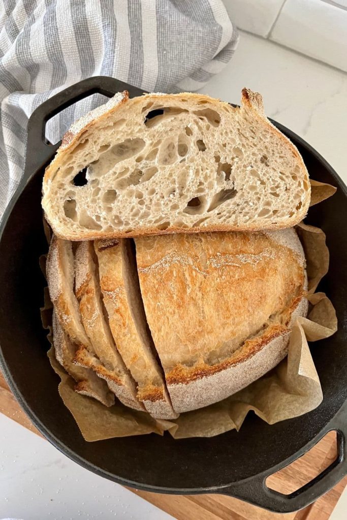 Loaf of sourdough bread sitting in a cast iron Dutch Oven. THe bread has a few slices sitting on top of the loaf showing the crumb.