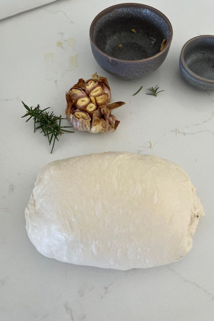 Sourdough dough that has been shaped into a batard. There is rosemary and roasted garlic inside the dough. There is a head of roasted garlic and some fresh rosemary above the bread.