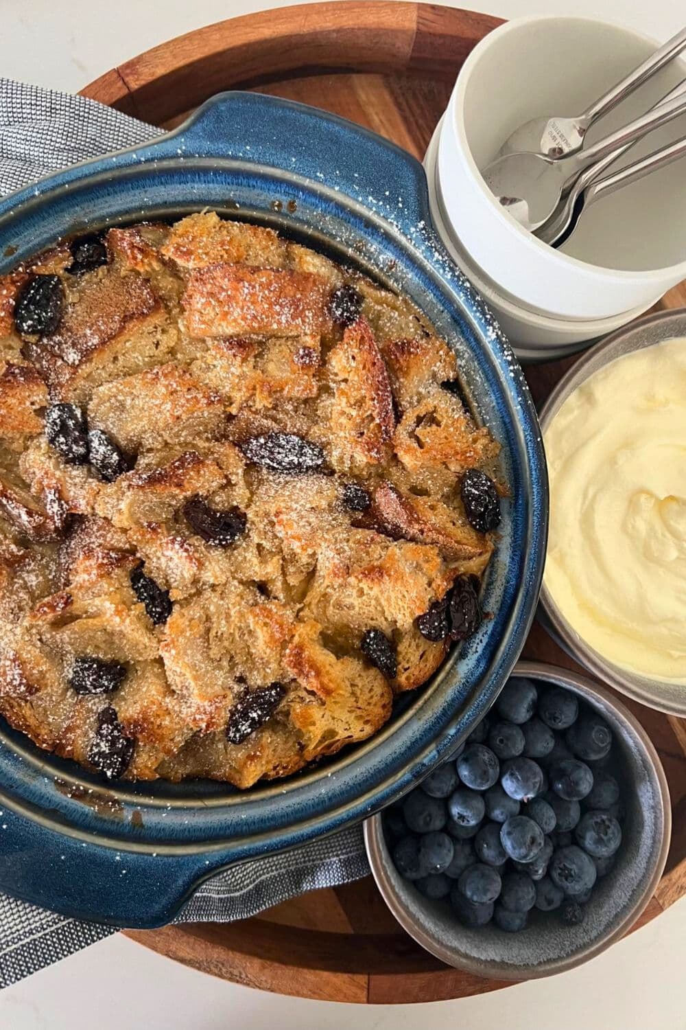 A sourdough bread pudding baked in a blue stoneware bowl. The pudding is surrounded by a bowl of fresh blueberries and a bowl of whipped cream.