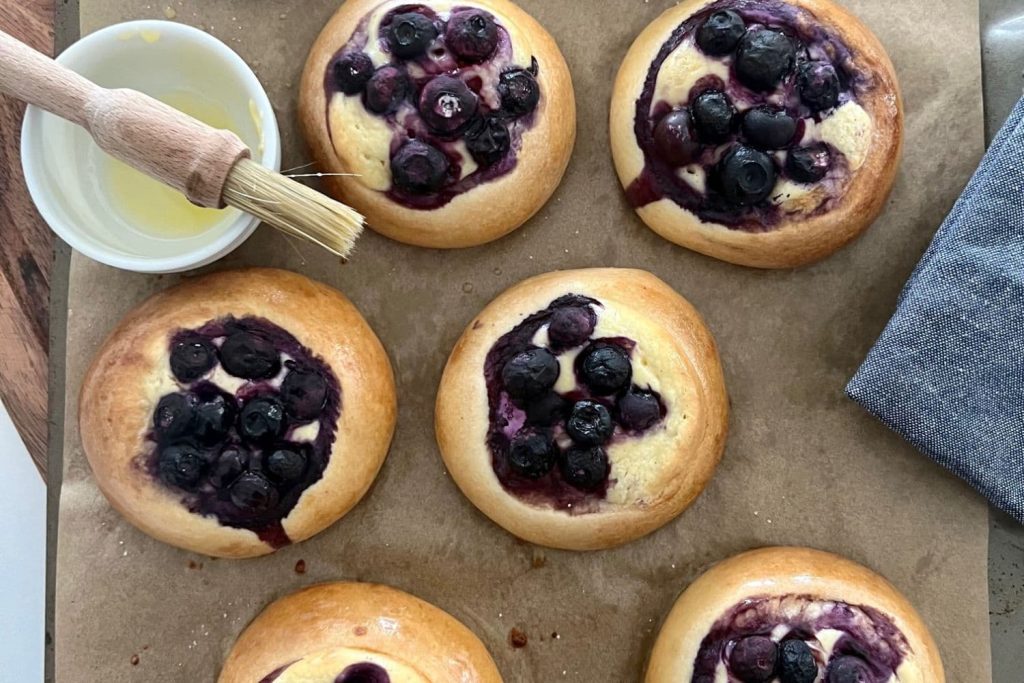 Sourdough Brioche Blueberry Cheesecake Rolls sitting on a piece of parchment paper with a pastry brush next to them.