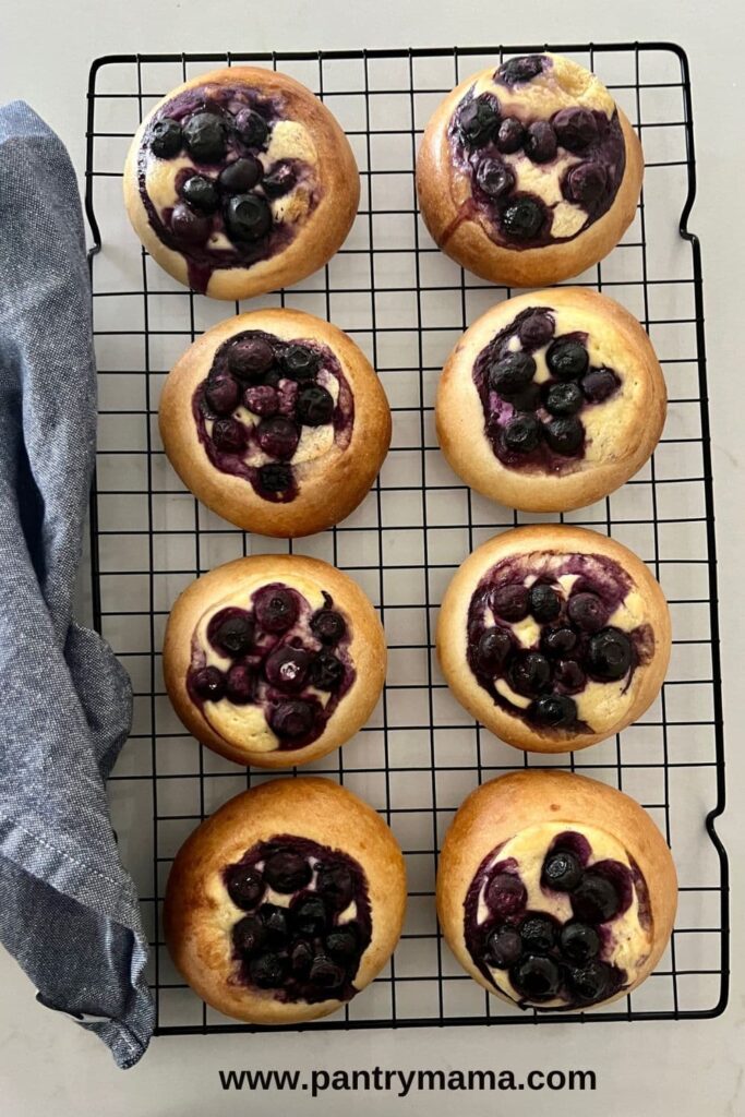 Sourdough brioche blueberry cheesecake rolls sitting on a wire rack with a blue dish towel sitting on the side.