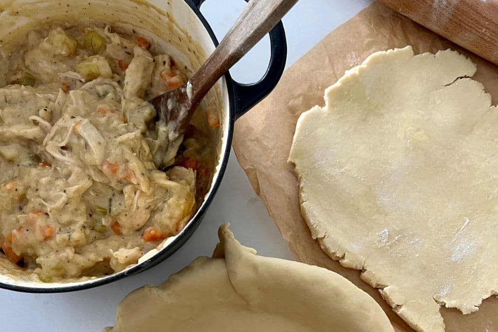 Sourdough chicken pot pie filling in a cream colored Dutch Oven. There is an unfilled pie crust next to the pot and some more pastry next to a rolling pin next to that.