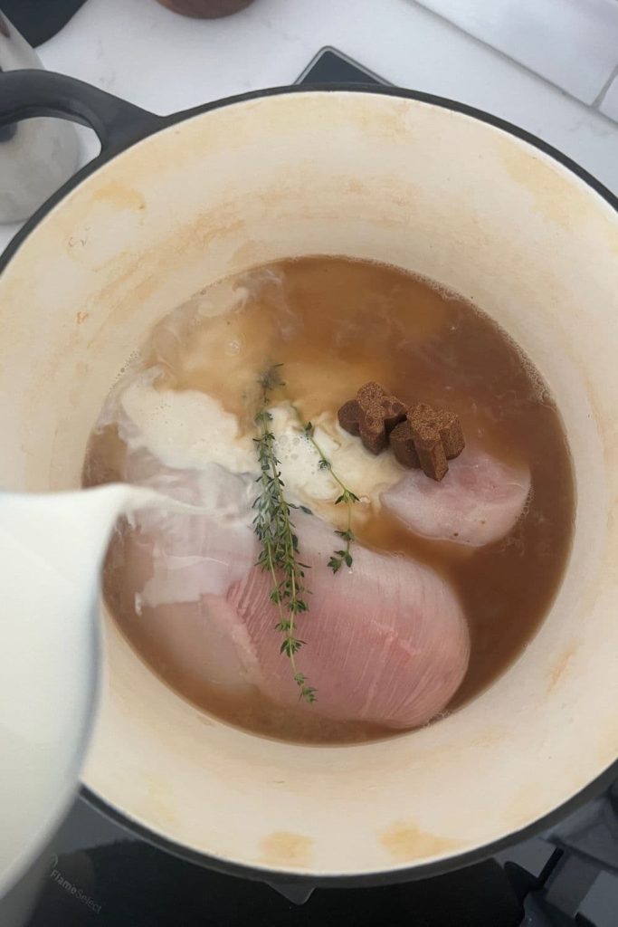 2 raw chicken breasts sitting in a heavy based pot. The chicken breasts are surrounded by chicken broth. There are thyme sprigs and stock cubes sitting on top. A jug of milk is being added to the pot.