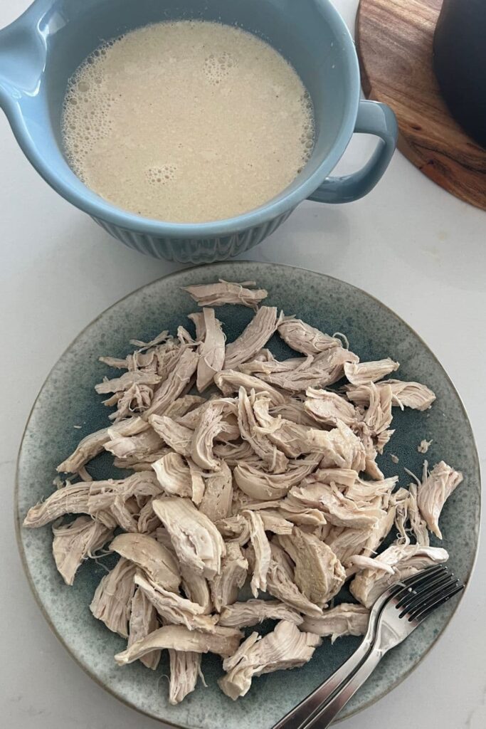 A blue mixing jug filled with the chicken poaching liquid is sitting next to a plate of shredded chicken.