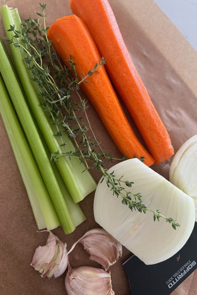 A wooden board with carrots, celery sticks, garlic cloves, white onions and thyme sprigs.