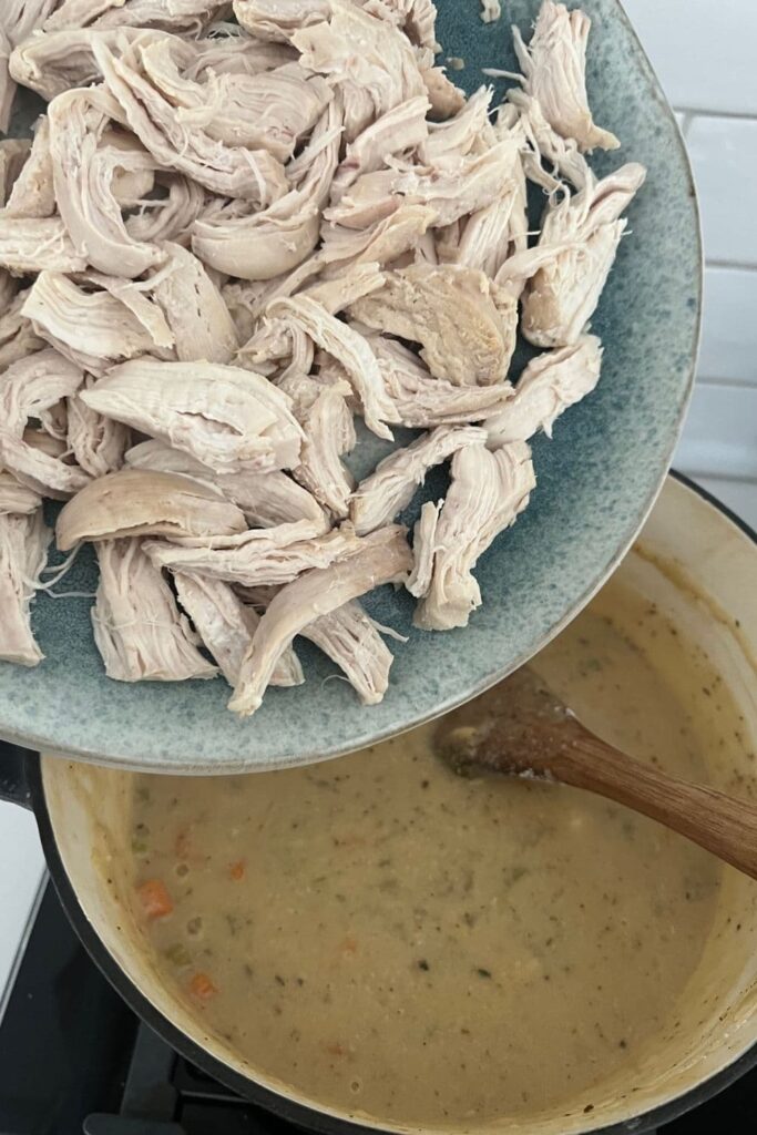 Heavy based pot with the creamy chicken pot pie vegetable sauce. The shredded chicken is being tipped into the creamy sauce.