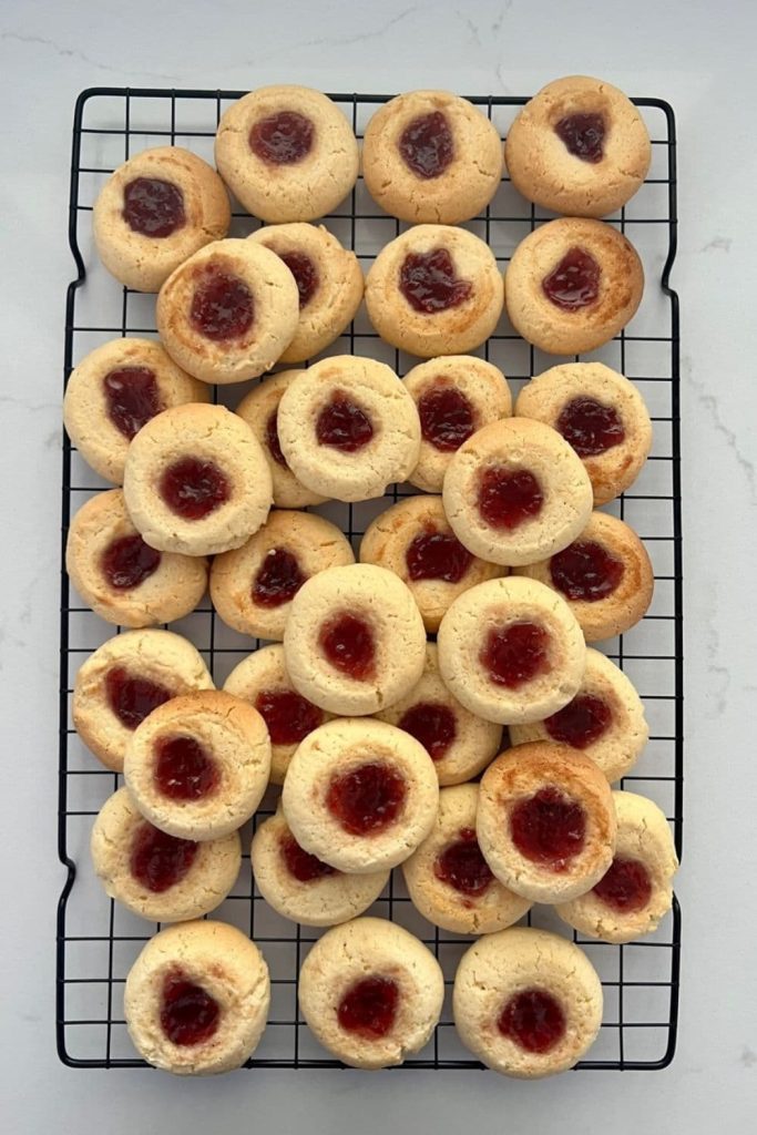 A stack of sourdough thumbprint cookies filled with strawberry jam sitting on a black wire cooling rack.