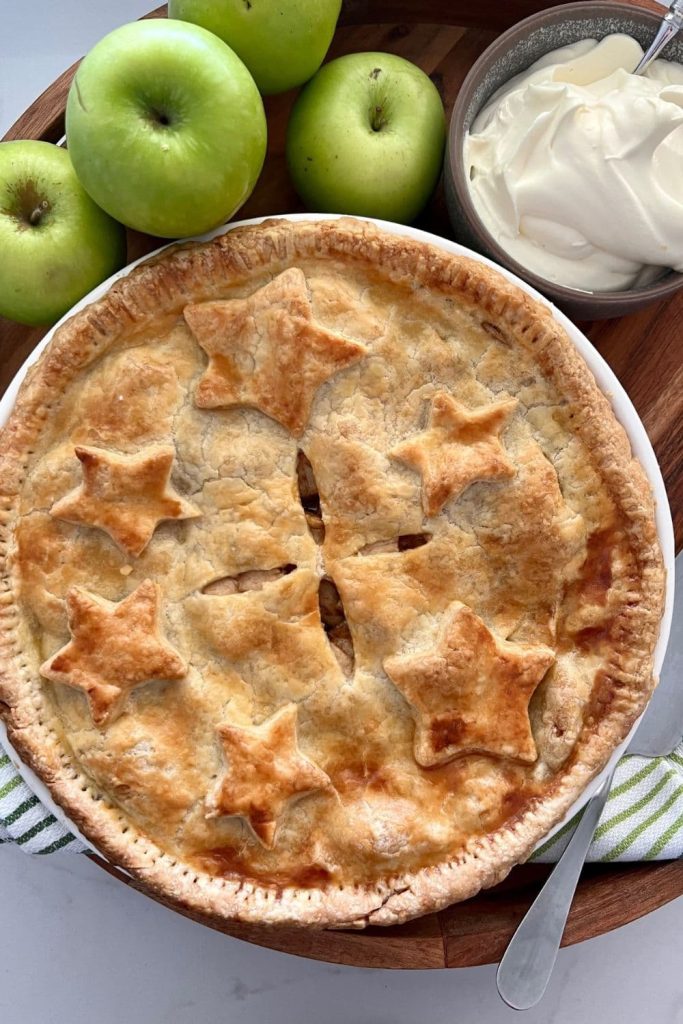 A whole sourdough apple pie shown with whole green apples and a bowl of whipped cream to complete the scene of sourdough comfort food.
