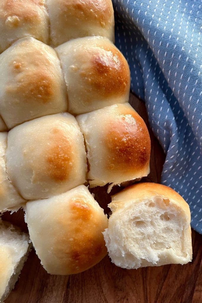 Soft sourdough dinner rolls on a wooden board. There is a light blue dish towel next to the rolls.