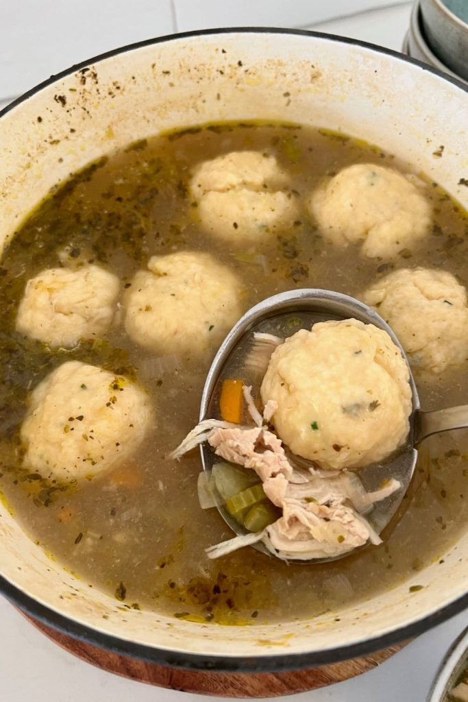 A heavy cream colored pot full of chicken soup and sourdough dumplings. Some of the chicken and dumplings are being ladeled out of the pot.