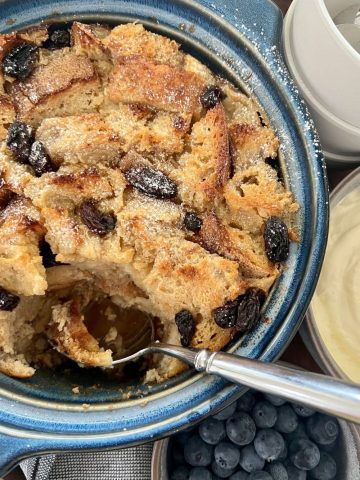 A blue casserole dish filled with sourdough bread pudding that is being scooped out with a silver spoon.