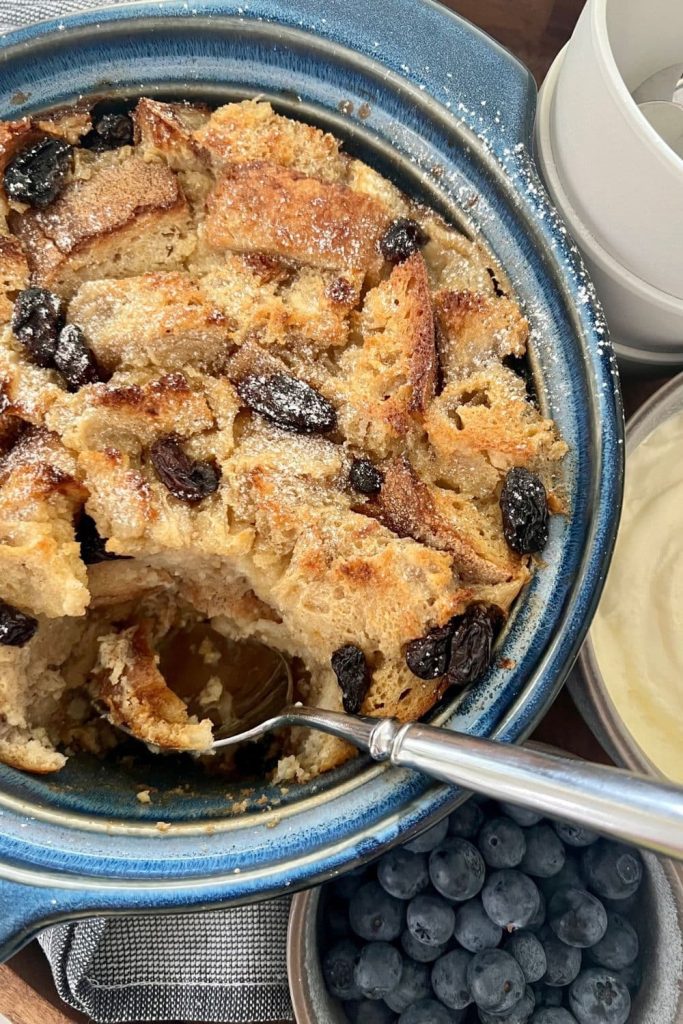 A blue casserole dish filled with sourdough bread pudding that is being scooped out with a silver spoon.
