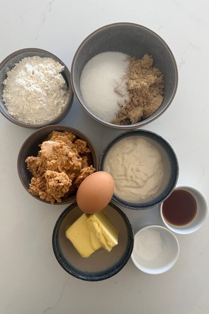 Several bowls containing the ingredients for sourdough discard peanut butter cookies - all purpose flour, white and brown sugar, peanut butter, egg, butter, vanilla extract, baking powder.