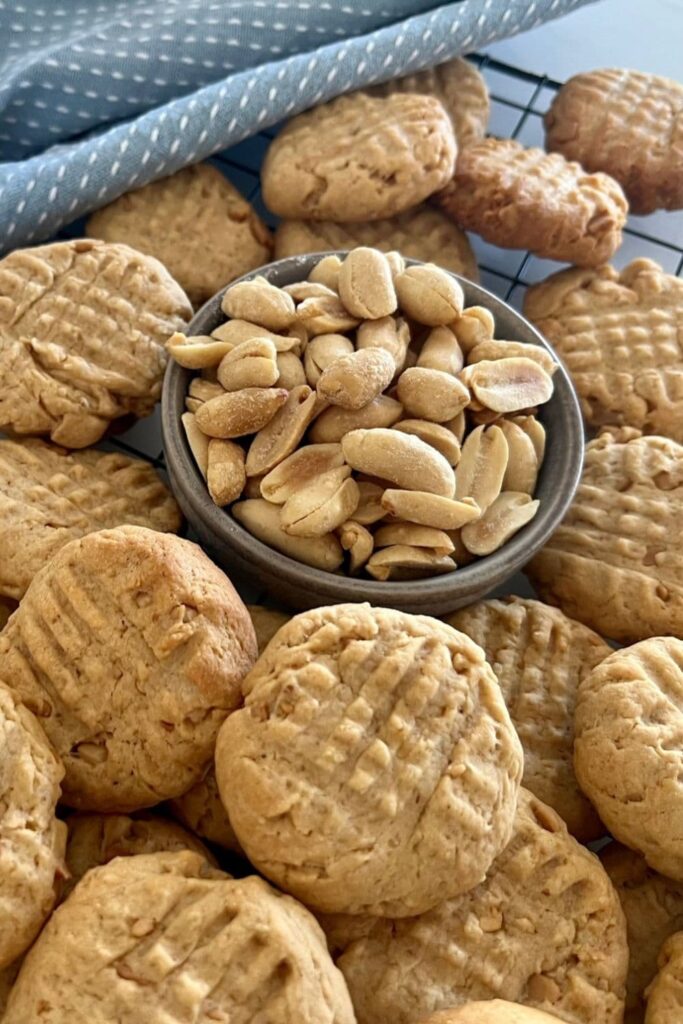 Sourdough peanut butter cookies piled up on a wire rack. There is a bowl of peanuts in the middle of the pile.