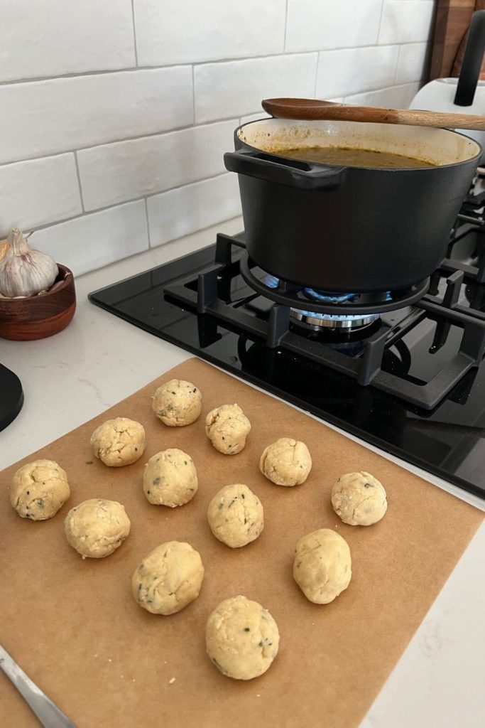 12 sourdough dumplings sitting on a piece of parchment paper. The dumplings are next to a black glass cooktop. There is a large black cast iron pot on the stove with simmering soup.