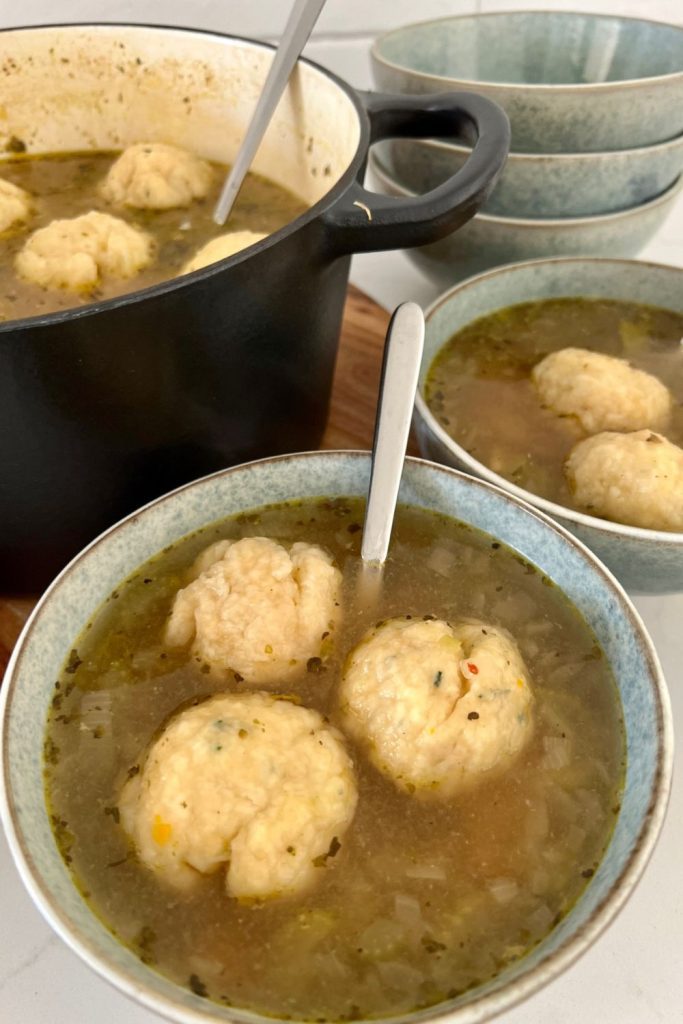 Two bowls of chicken soup with 3 sourdough dumplings in each bowl. There is a black Dutch oven in the background with the rest of the soup and dumplings in it.